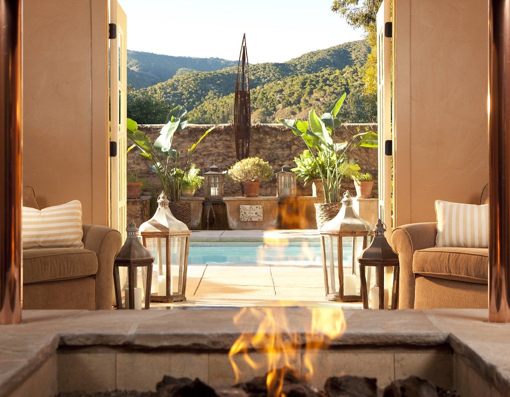 An elegant hotel suite with cream-colored linens, stone fireplace, and French doors opening to a private terrace with mountain views