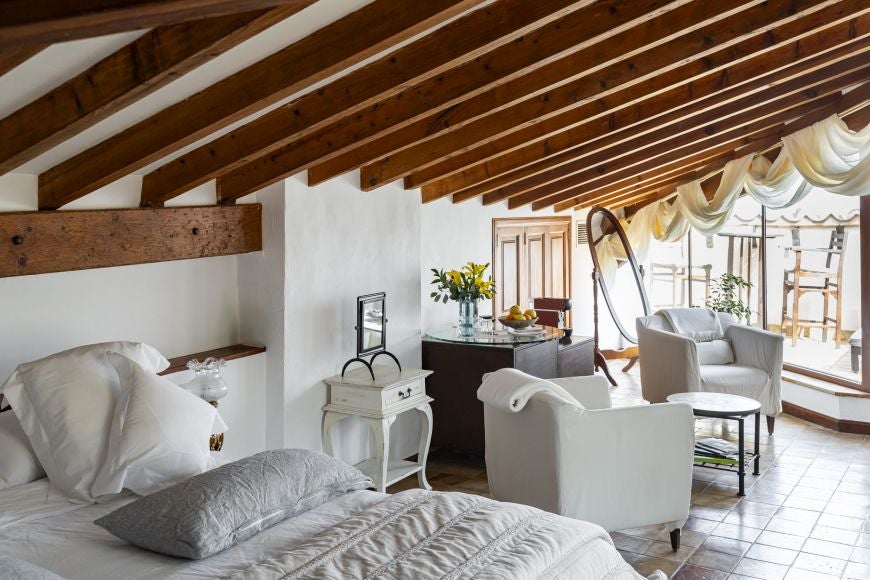 Elegant, sun-drenched deluxe room with rustic stone walls, plush white bedding, and panoramic views of Spanish countryside landscape