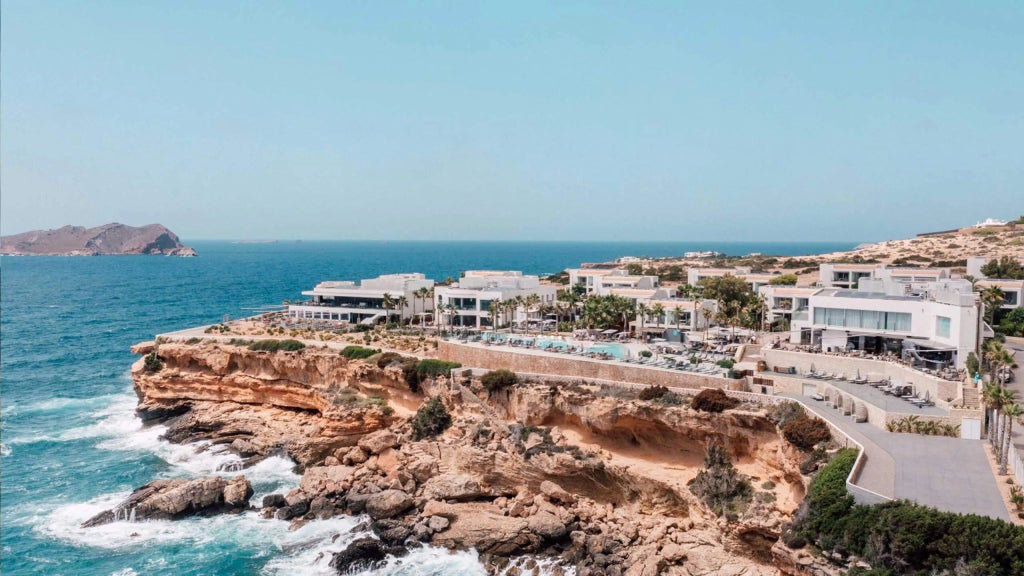 Luxurious white-walled resort overlooking azure Mediterranean waters, with infinity pool seamlessly blending into Ibiza's dramatic coastal landscape at sunset