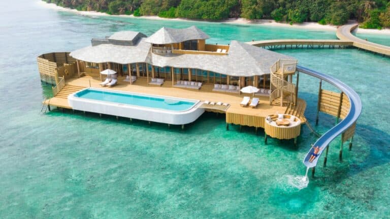 Overwater luxury villa with thatched roof, surrounded by turquoise lagoon and coral reefs in Maldives, viewed from aerial perspective