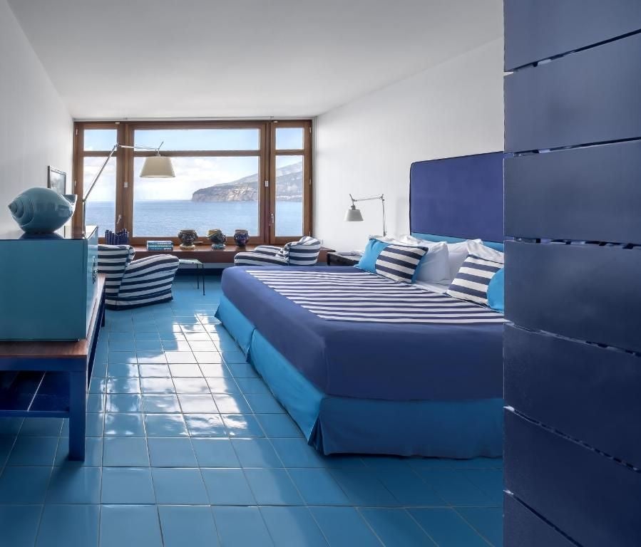 Luxurious Mediterranean hotel room with blue-white nautical decor, floor-to-ceiling windows overlooking Sorrento's coastline and bay