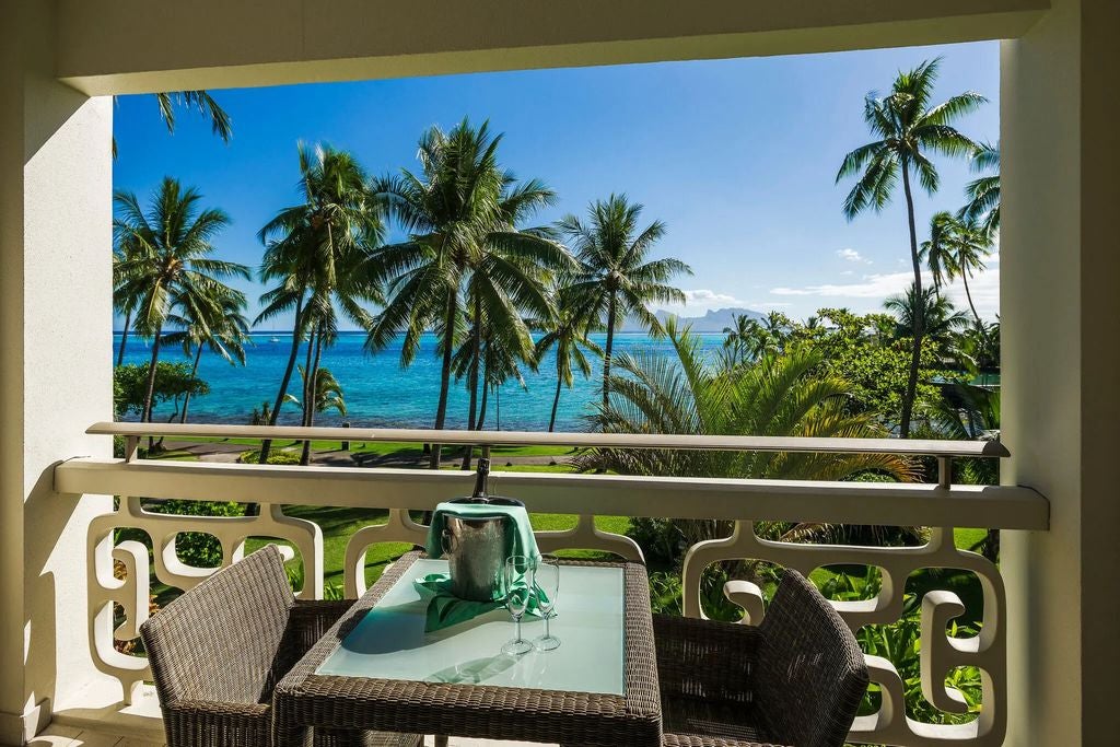 Elegant oceanfront hotel room with king bed, private balcony, teak wood furnishings, and tropical decor overlooking Tahitian lagoon