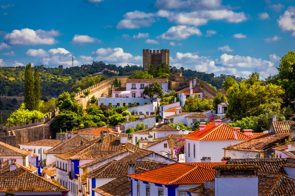 Scenic cobblestone street in Óbidos with whitewashed buildings, vibrant bougainvillea, and medieval castle walls framing historic Portuguese landscape