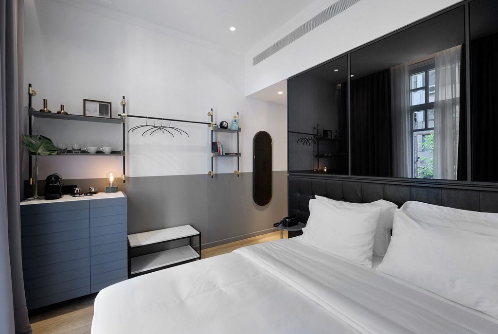 Minimalist hotel room with soft gray walls, crisp white linens, sleek wooden furniture, and large window overlooking urban Thessaloniki landscape.