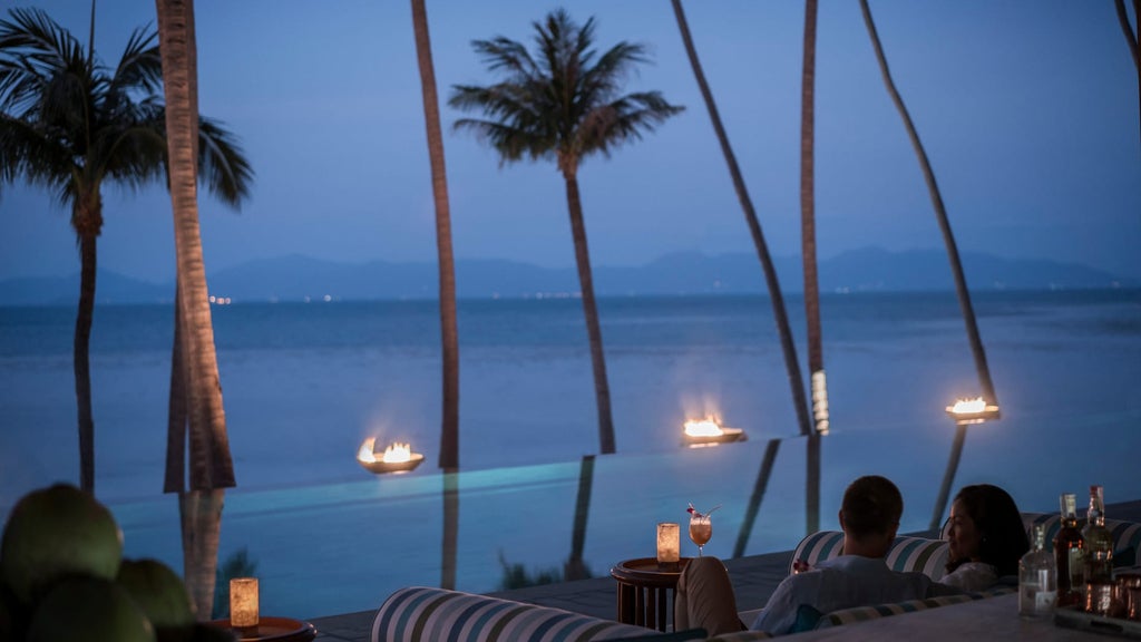 Oceanfront infinity pool surrounded by tropical palms overlooking Gulf of Thailand, with luxurious sun loungers and beach views