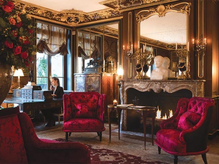 Opulent Parisian hotel exterior with ornate Haussmanian facade, wrought-iron balconies and burgundy awnings overlooking tree-lined avenue