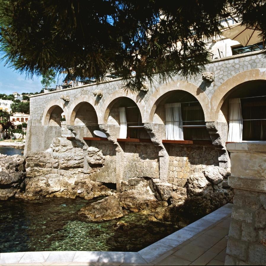 Elegant Mediterranean hotel with white stone facade, oceanfront terrace, and infinity pool overlooking dramatic rocky coastline at sunset