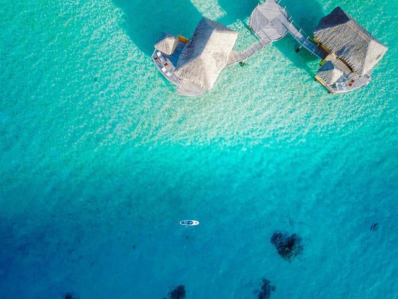 Luxurious overwater bungalows with thatched roofs stretch across turquoise lagoon waters, framed by lush palm trees and Mount Otemanu