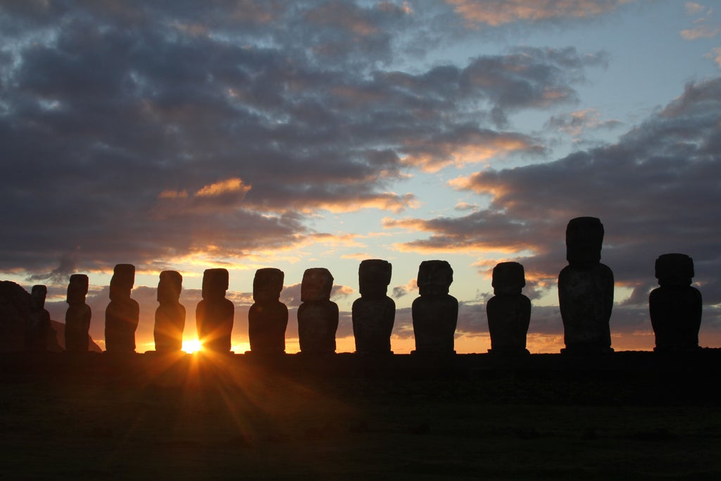 Iconic Easter Island moai statues silhouetted against vibrant orange sunset sky, with Pacific Ocean waves crashing at base of cliffs