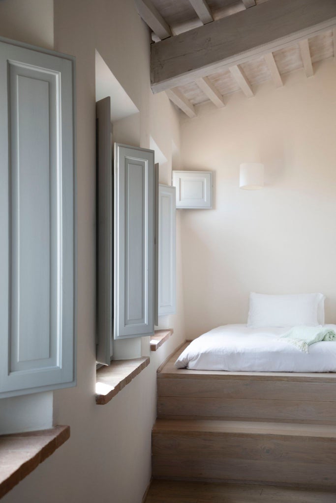 Elegant Italian boutique hotel room with neutral tones, minimalist design, plush white bedding, rustic stone wall, and warm natural lighting