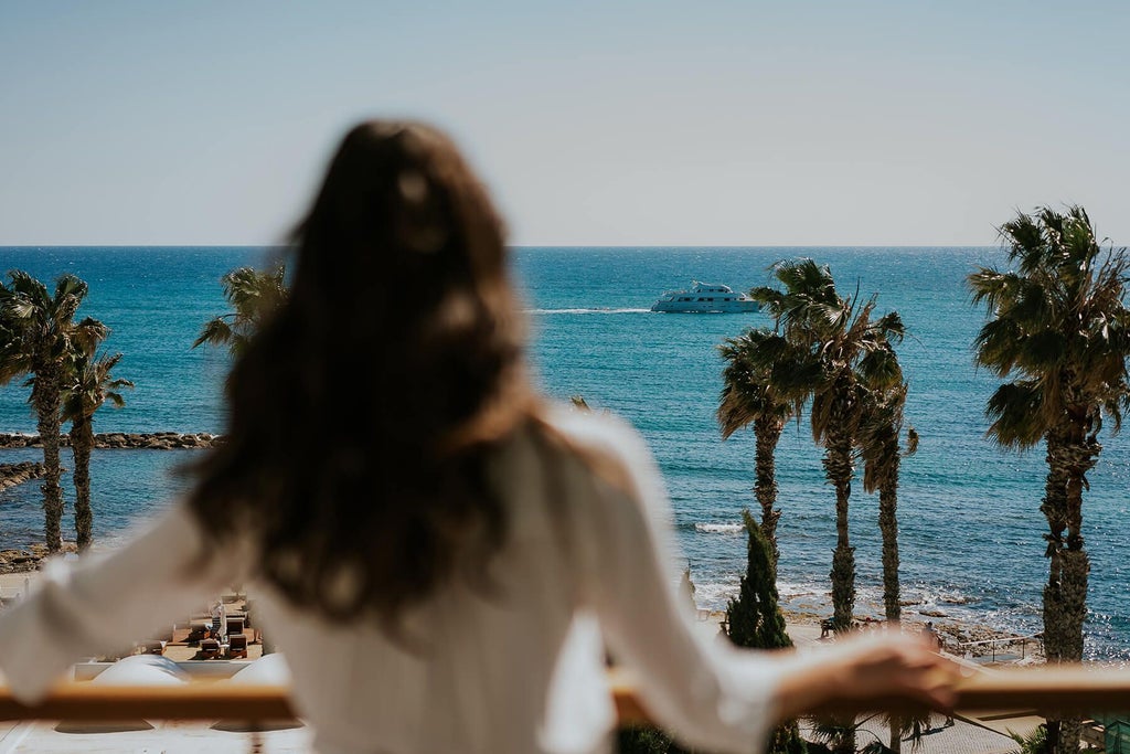 Luxurious beachfront hotel with modern white architecture, azure Mediterranean waters, and palm-lined tropical landscape in Cypriot resort setting.