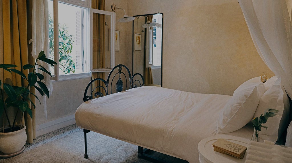 Elegant boutique hotel room with antique furnishings, soft neutral palette, ornate wooden bed, marble-topped side tables, and soft Greek sunlight filtering through sheer curtains.