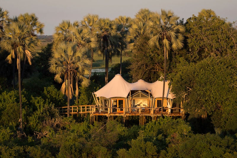 Elevated luxury safari lodge with thatched roof cottages connected by wooden walkways, overlooking vast Okavango Delta wetlands