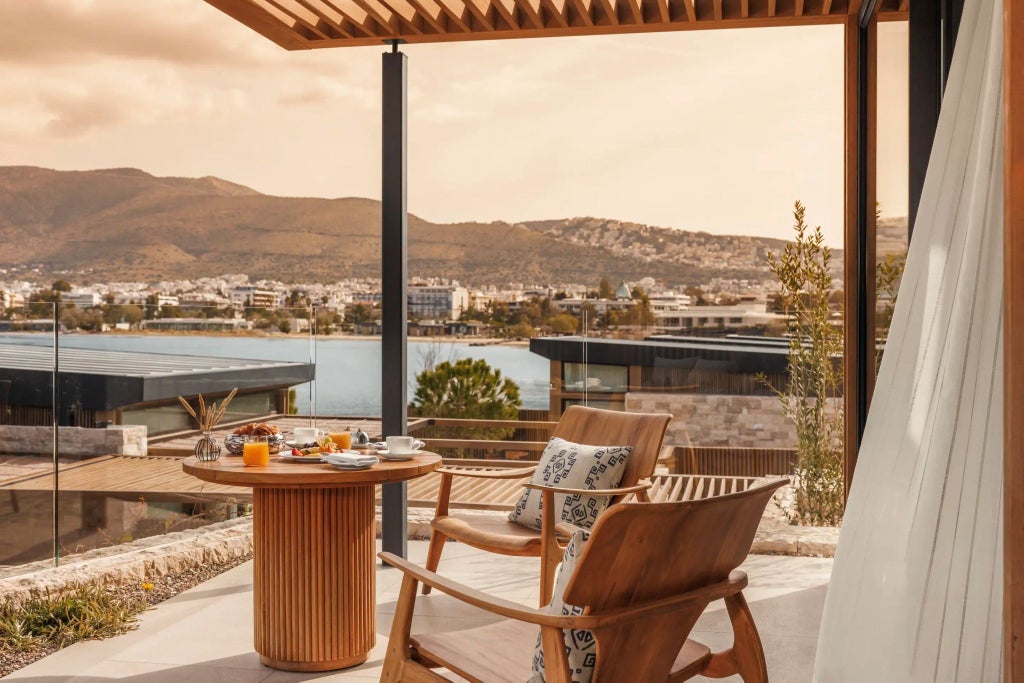 Sunlit luxury bungalow with minimalist design, wooden floors, panoramic windows overlooking Grecian coastline, elegant white furnishings at One&Only Aesthesis resort