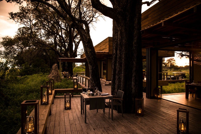 Elevated luxury safari lodge with thatched-roof pavilions connected by wooden walkways overlooking a pristine Botswana floodplain