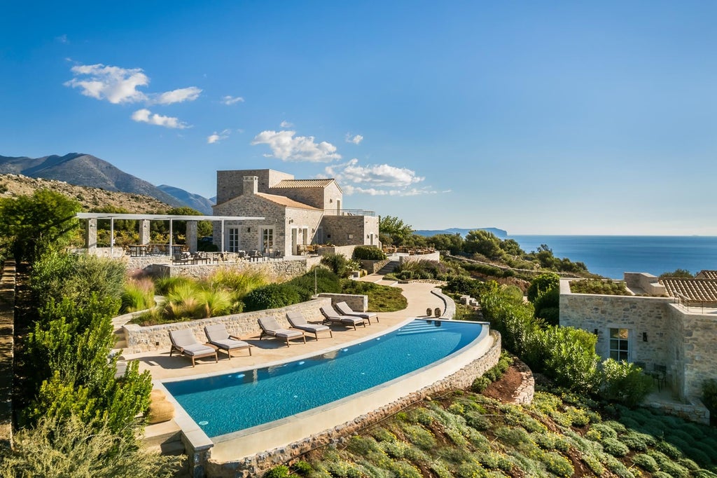 Elegant stone Mediterranean villa with terracotta roof, nestled among olive trees, overlooking azure Aegean Sea with sunlit stone terrace and lush greenery