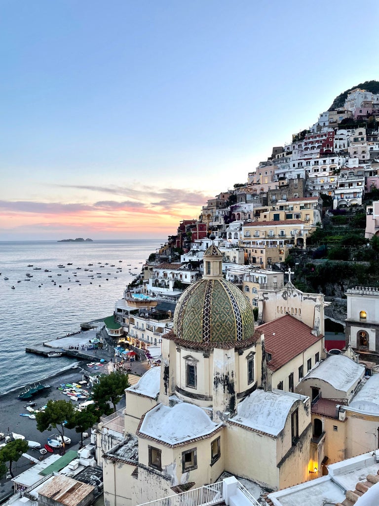 Blue wooden boat glides past dramatic Amalfi Coast cliffs, with colorful hillside buildings and crystal Mediterranean waters beneath