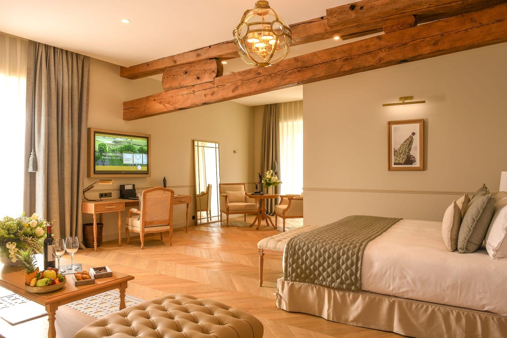 Elegant junior suite with plush white bedding, rich wood furniture, soft neutral tones, and large windows overlooking French cognac estate architecture