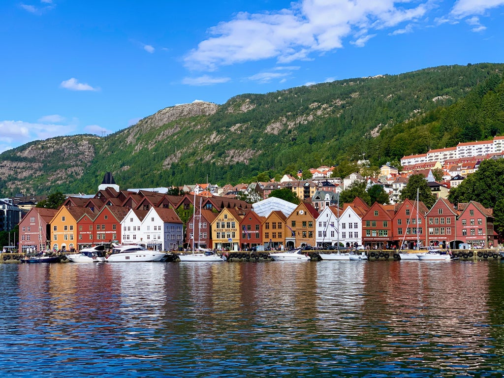 Snow-capped mountains surround Bergen's historic harbor, featuring colorful wooden Hanseatic buildings and luxury yachts along the waterfront