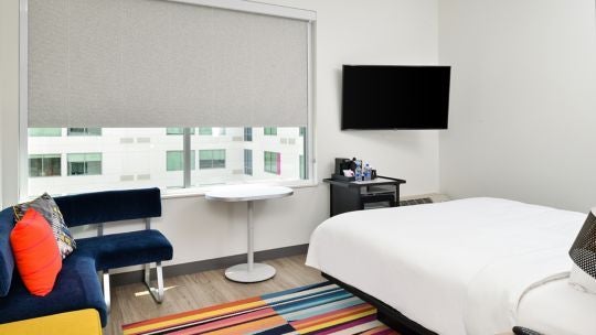 Modern, spacious Aloft hotel king room with sleek contemporary design, crisp white linens, minimalist furniture, and city skyline view through large windows.