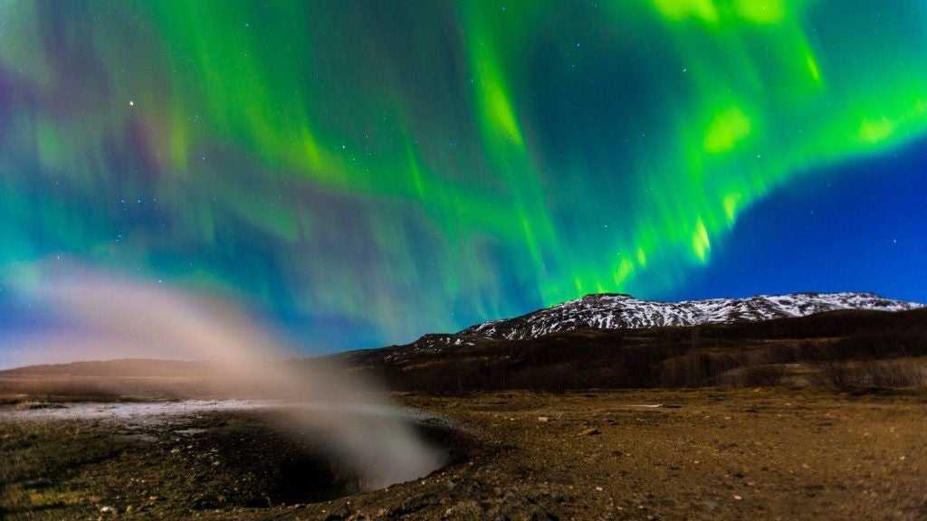 Rustic wooden hotel exterior with black roof overlooking dramatic Icelandic geothermal landscape, featuring vibrant green moss and distant steaming geysers at dusk