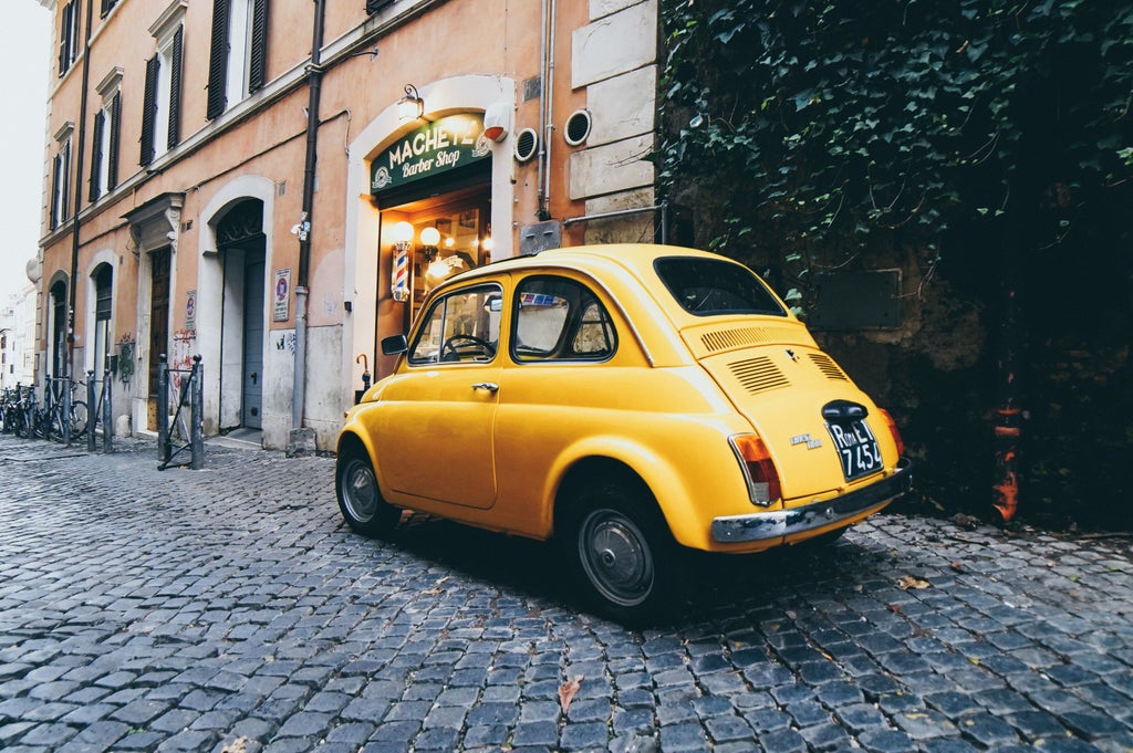 Vintage red Fiat 500 car parked in narrow cobblestone street with rustic Italian buildings and charming window shutters in Florence