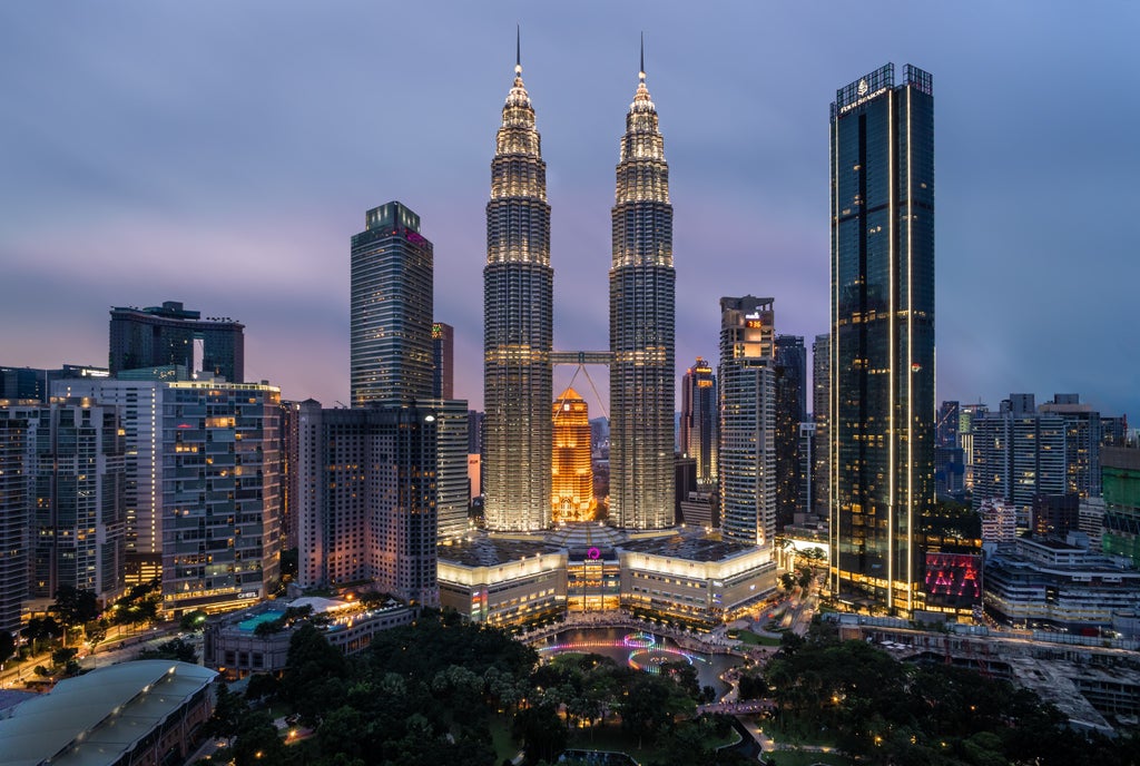 Illuminated Petronas Twin Towers dominate KL's night skyline, reflecting on lake with modern skyscrapers and lush gardens below