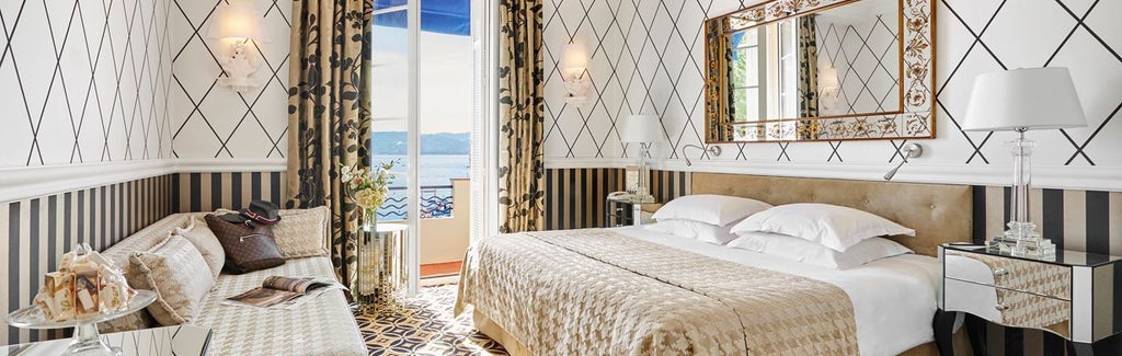 Elegant hotel room with panoramic Mediterranean views, featuring rich wooden furnishings, king bed, and opulent French Riviera decor