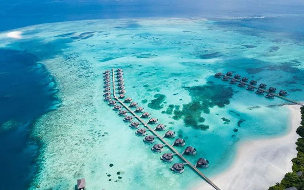 Luxurious overwater villa with wooden walkway in Maldives, surrounded by turquoise lagoon and swaying palm trees at sunset.