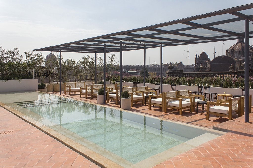 Elegant minimalist hotel courtyard in Mexico City, with neutral stone walls, geometric lines, modern furniture, and lush greenery casting dramatic shadows