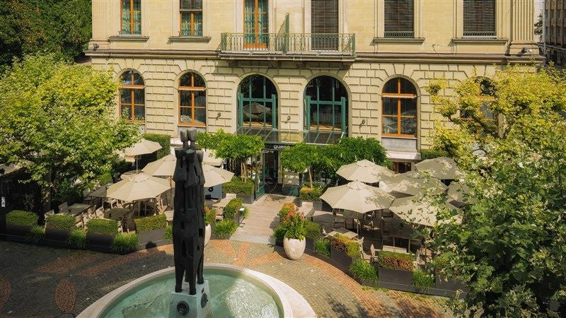 A grand lakefront luxury hotel with Belle Époque facade, ornate balconies and iconic red awnings overlooking Lake Geneva at sunset