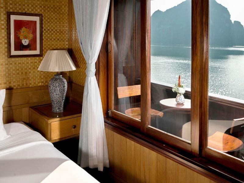 Spacious premium balcony cabin on Bhaya Cruises with elegant wood furnishings, panoramic windows overlooking scenic Halong Bay waters, creating a luxurious maritime retreat