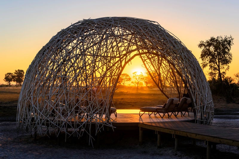 Elevated luxury safari lodge with thatched-roof villas on stilts overlooking lush Okavango Delta wetlands at golden hour