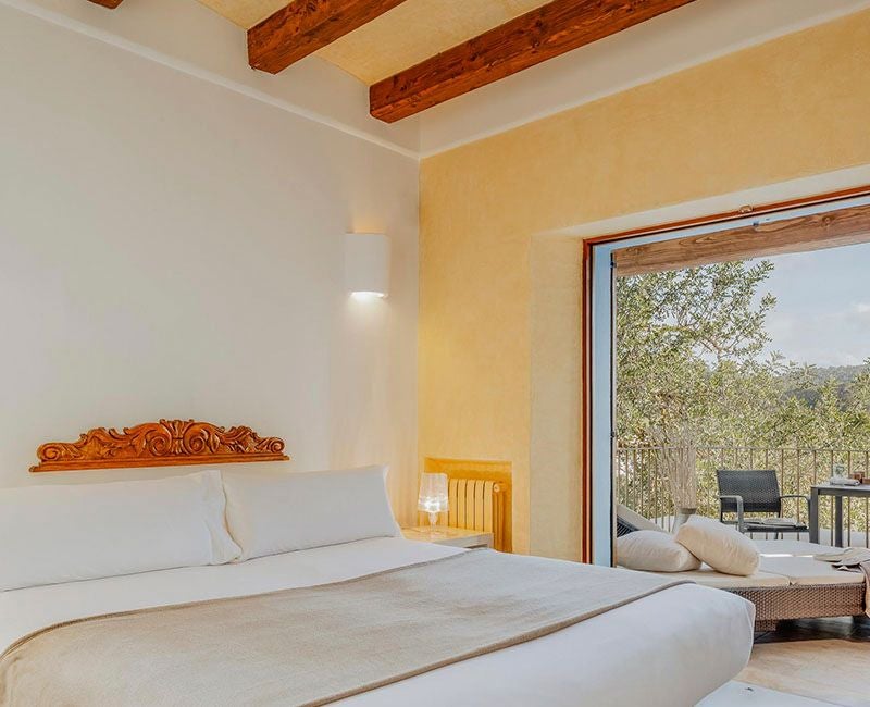 Elegant white-walled hotel room with minimalist design, wooden floor, crisp white bed linens, and natural light streaming through large windows at Ca Na Xica.