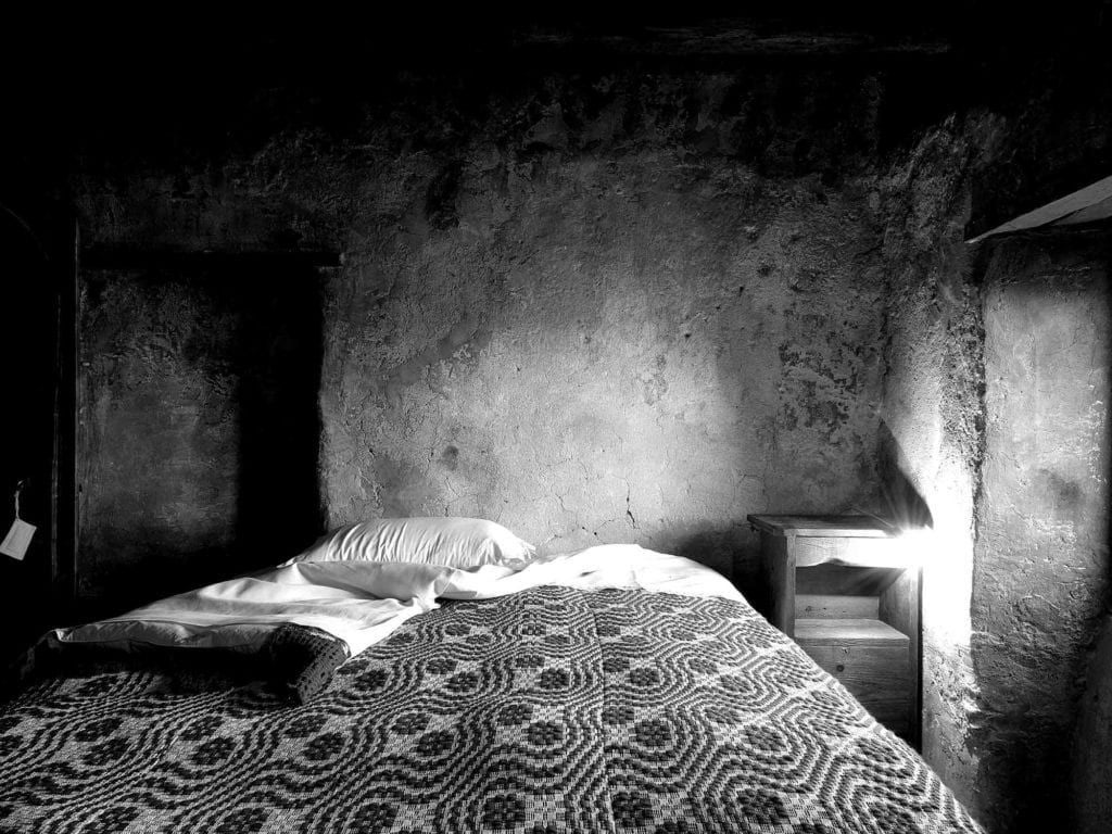 Rustic stone-walled hotel room with minimalist wooden bed, soft neutral linens, and traditional Italian architectural details highlighting historic charm