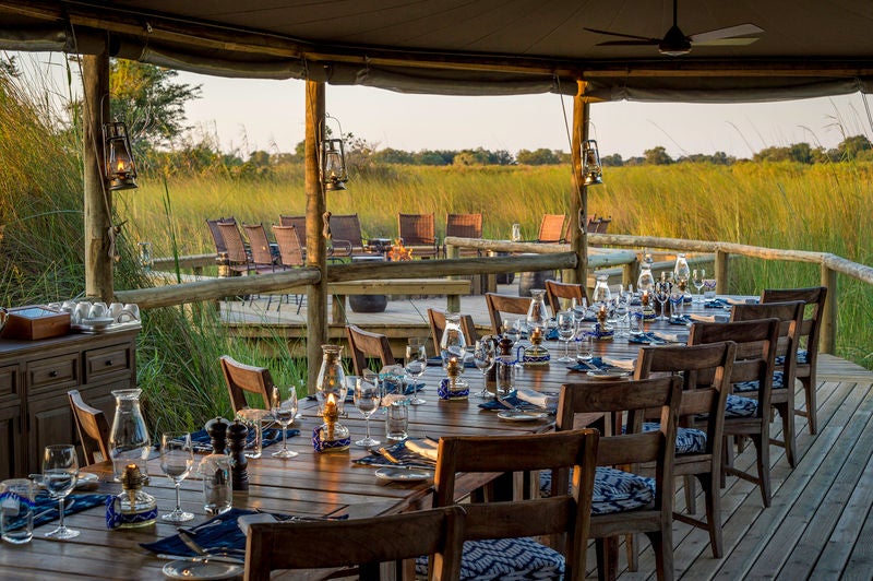 Luxury safari lodge with thatched-roof suites elevated on wooden platforms, overlooking flooded Okavango Delta grasslands at sunset