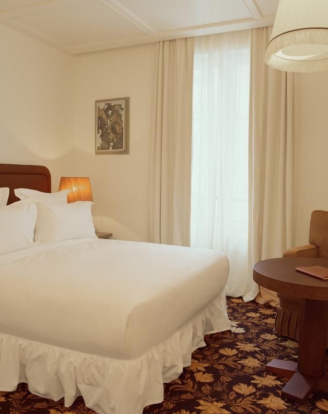 Elegant Parisian hotel room with plush king bed, cream-colored walls, ornate chandelier, and large arched window overlooking city streetscape