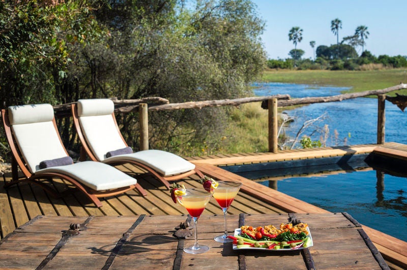 Elevated luxury safari tent with wooden deck overlooking wetlands, surrounded by lush acacia trees in Okavango Delta wilderness