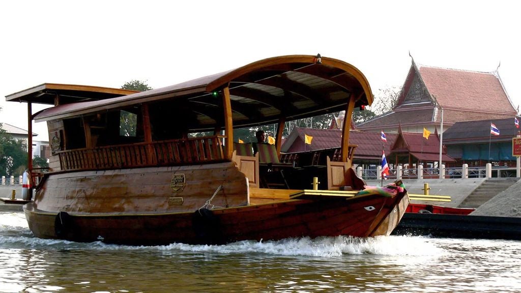 Ancient Ayutthaya temple ruins viewed from a luxurious tuk tuk, with a traditional river cruise boat gliding past historic Buddhist architecture