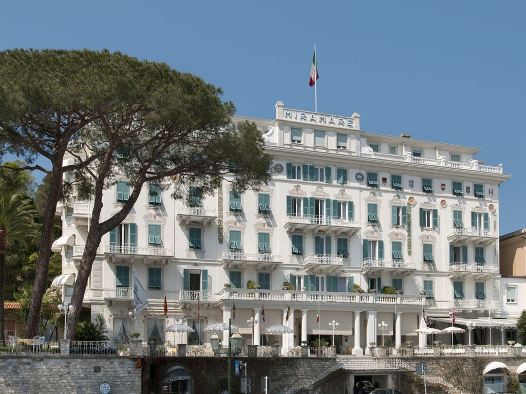 Elegant white Mediterranean hotel with ornate balconies overlooks Italian Riviera coastline, palm trees frame manicured garden terrace