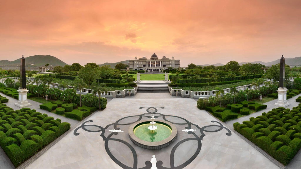 Elegant white palace-style resort with ornate domes and archways reflected in Lake Udai Sagar, surrounded by mountains at sunset