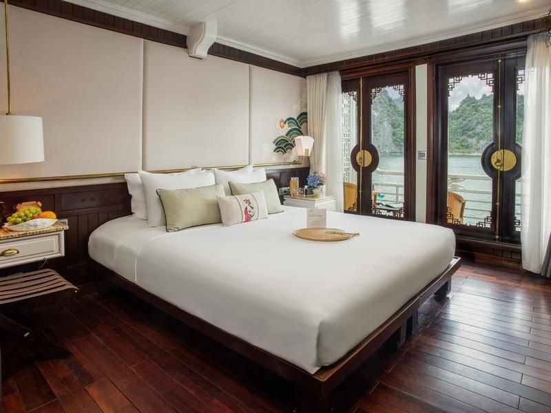 Luxurious Bhaya Cruises deluxe cabin with hardwood furnishings, plush bed, elegant Vietnamese decor, and large window overlooking emerald Halong Bay waters