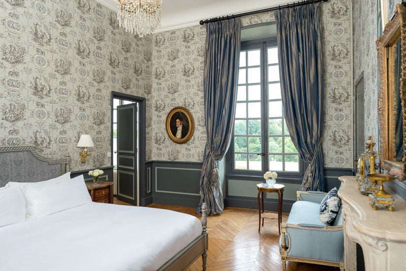 Elegant French château hotel room with ornate period furniture, crystal chandelier, antique mirrors and garden views through tall windows