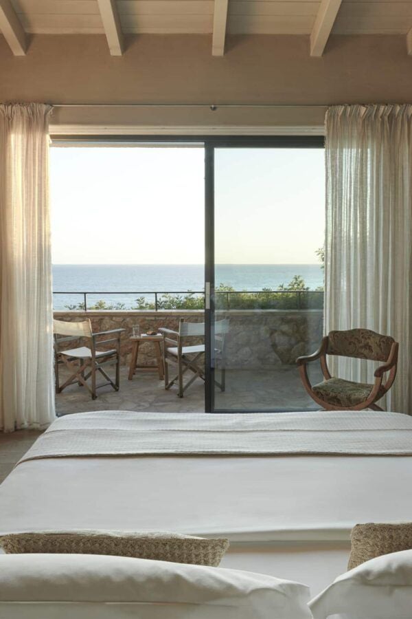 Minimalist white hotel room with panoramic sea view, elegant wooden furniture, and large windows showcasing Kefalonia's coastline at sunset