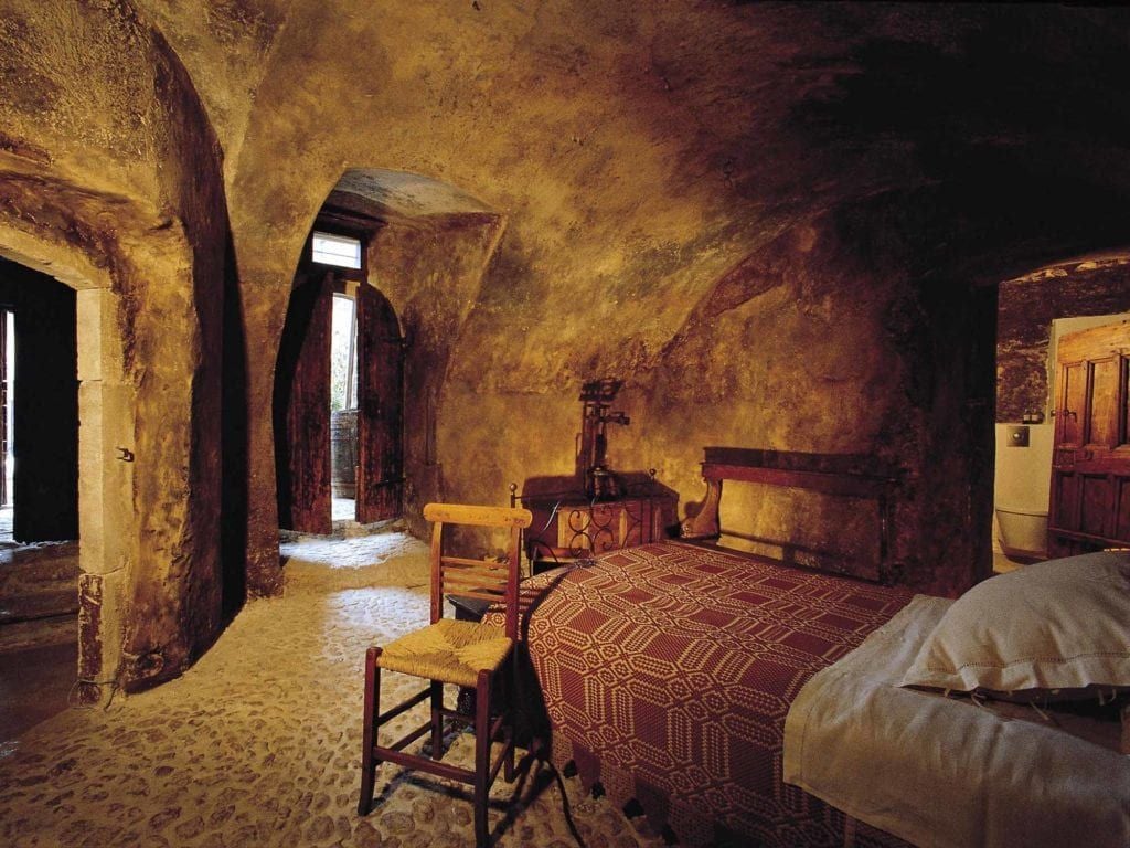 Rustic Italian stone room with antique wooden bed, arched window overlooking historic village, soft natural light illuminating traditional architectural details