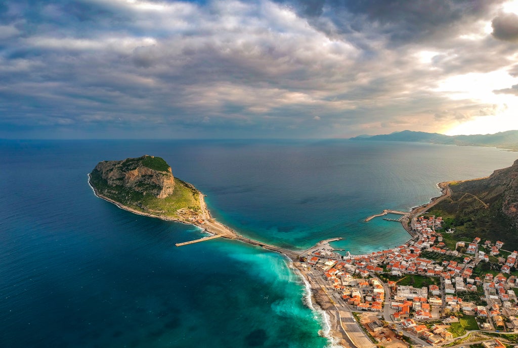 Luxurious stone-walled medieval town of Monemvasia perched on rocky coastal cliff, golden sunlight illuminating historic Byzantine architecture and azure Aegean Sea backdrop