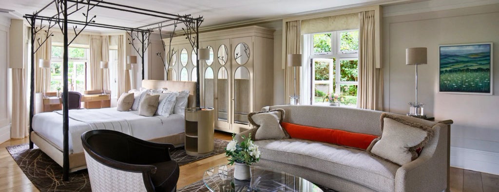 Elegant bedroom in Gardener's East Cottage Suite at Coworth Park, featuring plush white bedding, rustic wooden furniture, and soft natural lighting.