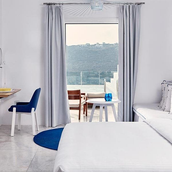 Luxurious white-themed hotel room with panoramic Aegean Sea view, minimalist design, private balcony, and elegant blue accents at Myconian Ambassador Hotel