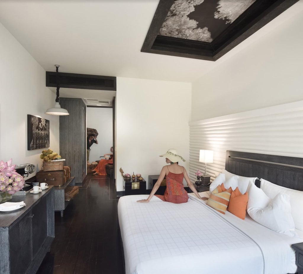 Hotel suite with elegant dark wood furnishings, crisp white bedding on king bed, ambient lighting, and Khmer-inspired wall decor