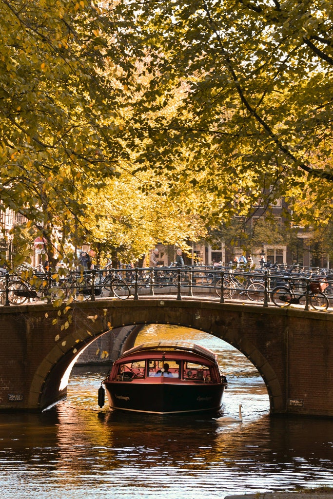 Elegant wooden boat glides through Amsterdam's scenic canals at sunset, passing historic canal houses with ornate facades and bicycles